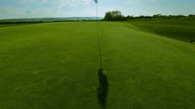 Pan shot of a green golf course, hole and flag on a bright sunny day. Sport, relax, recreation and leisure concept. Summer landscape with sunbeams in the sky