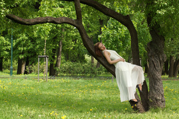 A beautiful girl in a long white dress leans against a tree branch in a summer park