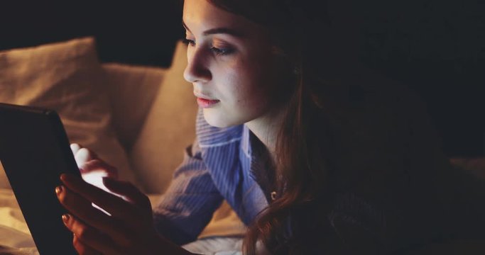 Woman using Tablet Computer at night. SLOW MOTION 4K DCi. Young millennial consumer shopping online from home, lying on bed. Technology in everyday life. 