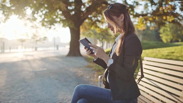Young Businesswoman Using Digital Tablet, Sitting on the Bench in the Park in the Sunny Morning City. SLOW MOTION. STEADICAM Stabilized Shot. Business Woman Relaxing Outdoors. Lens Flare. 
