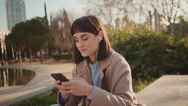 Young attractive cute millennial hipster woman chats and flirts on messenger application on smartphone while sitting outside in park in winter,concept social media, sexting and communication