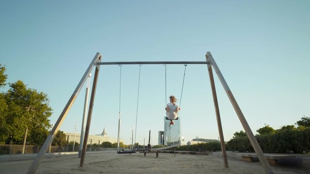 Lonely young woman in white tshirt and jeans swings on swingset on kids playground, flies high in air overlooking city skyline, concept forever youth and happinness