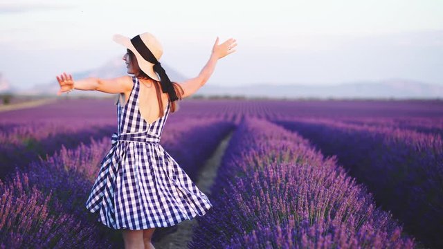 Happy Young Woman Enjoying Life in Lavender Field at Sunset. 4K. Joyful girl in a summer dress and straw hat dancing in endless blooming lavender fields.  Plateau du Valensole, Provence, France.
