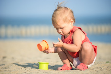 Baby playing on the sandy beach near the sea. Cute little girl in red dress with sand on tropical beach. Ocean coast.