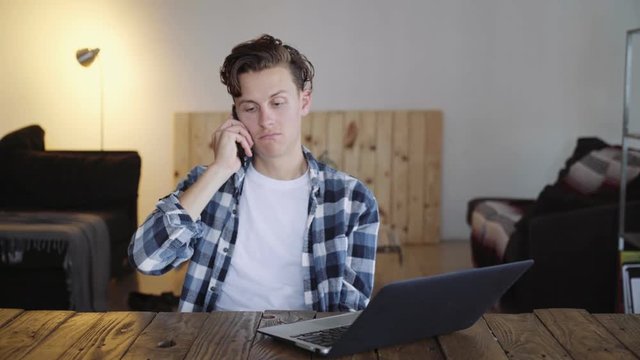 Handsome young man sits in coworking office, picks up smartphone to answer call, discusses urgent important matter, but gets angry fast and shouts at someone on other line