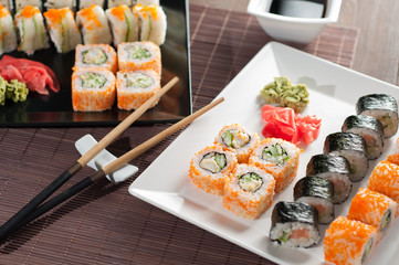 Sushi on a black and white plate, on a wooden mat with chopsticks and soy sauce