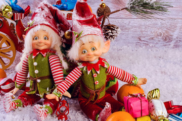 Funny christmas elves sits next to Christmas sock on light wooden background. Near tangerines and gifts.
