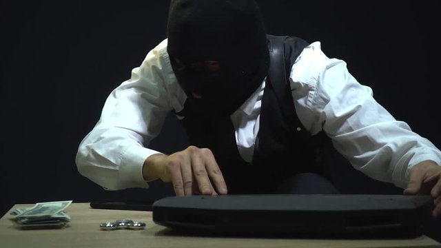 The robber in the mask examines a smartphone, a laptop, a bundle of dollars, takes a spinner from walks with him