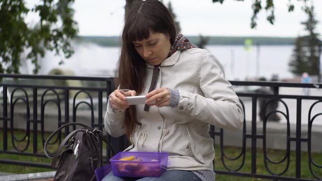Young brunette woman in a warm parka opens a container with food, then takes a photograph of a sandwich using a camera on a smartphone, sitting on a bench in a park next to a pond on a cloudy day