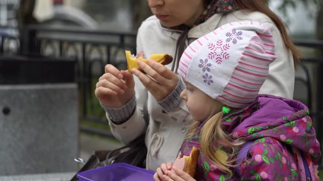 Mother and daughter eating sandwiches sitting on a bench in a cold overcast afternoon. Young family at lunch outdoors. A brunette woman and a little girl are dressed in warm outerwear.