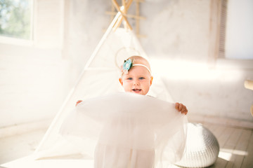 little girl one year with blue eyes blonde in a lush white dress is pleased and plays in a bright room on background of an Indian tent