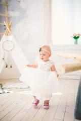 little girl one year with blue eyes blonde in a lush white dress is pleased and plays in a bright room on background of an Indian tent