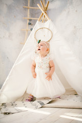 Fototapeta na wymiar little girl one year with blue eyes blonde in a lush white dress is pleased and plays in a bright room on background of an Indian tent