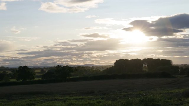 Rural autumnal landscape with brown mowed wheat field and sun setting over horizon, Tarvin, Cheshire, England, UK. Overcast sky with mountains and trees in background. Sunset