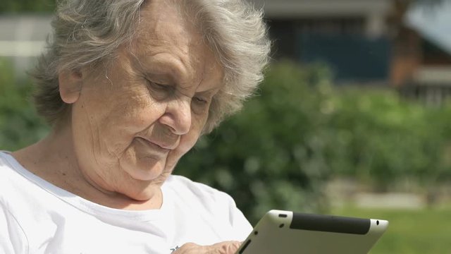 Mature elderly woman dressed t-shirt sitting on the background the green park outdoors in summer holds a silver portable computer tablet for looking the images. Slow motion
