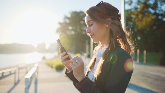 Young Businesswoman Using SmartPhone, Going to Work in the Sunny Morning City. SLOW MOTION. STEADICAM Stabilized Shot. Attractive Professional Business Woman with a cell phone in a city. Lens Flare. 