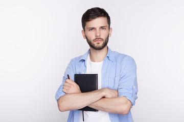 Fototapeta na wymiar Headshot of attractive serious European student with beard, mustache, dressed in casual t-shirt, looking at camera with indifferent stony facial expression, keeping his arms folded white studio wall