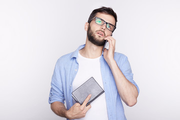 Handsome European young male freelancer in blue jacket talking on mobile while standing against white wall indoors, looking tired. Attractive hipster man having phone conversation during work at home.