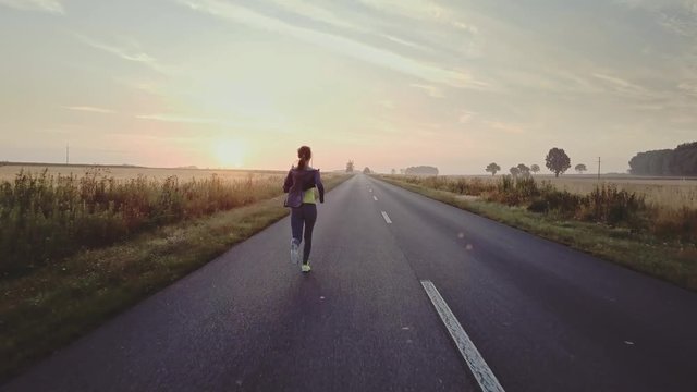 AERIAL 4K: Woman Running on a Countryside Road at Sunrise. Drone shot of a Female jogger athlete training at dusk. Active healthy lifestyle concept. Cinematic morning sports footage. 