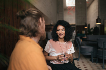 Beautiful smiling girl sitting in restaurant with friend. Pretty African American lady sitting at cafe with glass of water in hand. Young girl with dark curly hair talking with friend at cafe