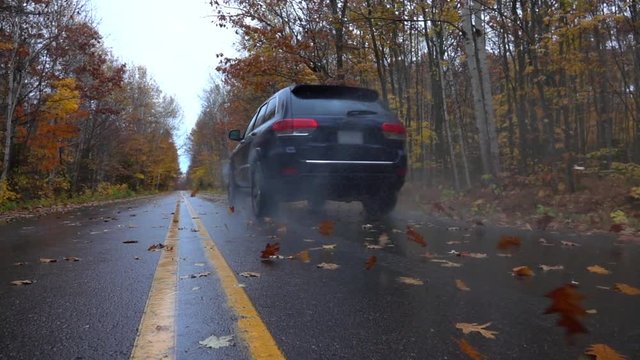 SLOW MOTION CLOSEUP: Black SUV car driving along an empty forest road, over fallen autumn tree leaves in reainy fall. Black jeep car driving fast along the wet slippery road, swirling colorful leaves