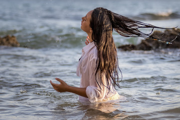 Chica joven con camisa blanca bañándose en el mar al atardecer y jugando con su pelo largo moreno.