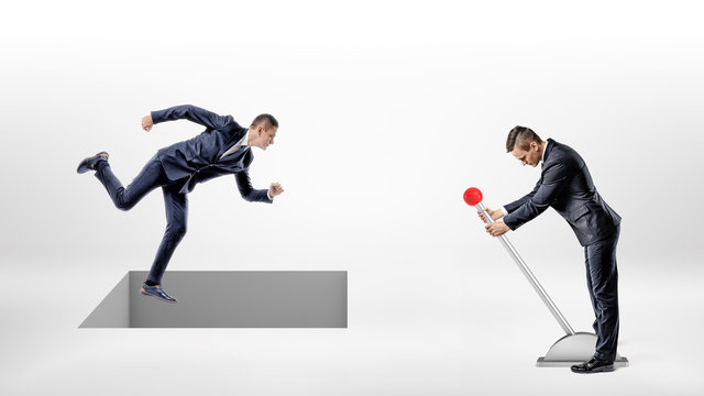 A businessman runs on white ground while a square hole controlled by a man on a lever opens right under his feet.