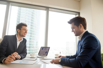 Satisfied businessman signing contract document while sitting at meeting desk in front of happy smiling business partner. Investor accepting agreement after studying project financial indicators