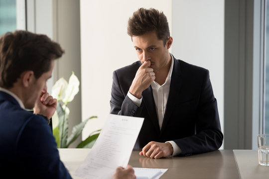 Nervous male job candidate looking with worriment on human resources generalist reading his resume during interview in company office. Millennial man trying to find job, giving application for vacancy