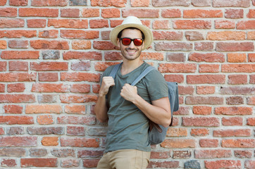 Outdoor photo of young hipster guy standing against wall of red bricks with gray backpack, wearing casual neutral clothes, sunhat and trendy sunglasses, looking at street waiting for city adventures