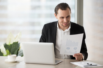 Young cheerful business man sitting in modern office in front of laptop with happy smile, holding financial report document, pleased with successful stock investment profit. Finance success concept.