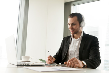 Fototapeta na wymiar Smiling businessman in headphones taking notes in front of laptop at workplace. Friendly young manager participating in online meeting or conference, remote job interview, learning foreign languages.