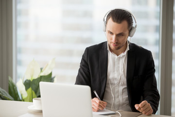 Thoughtful businessman in headphones taking notes in front of laptop at workplace. Young manager...