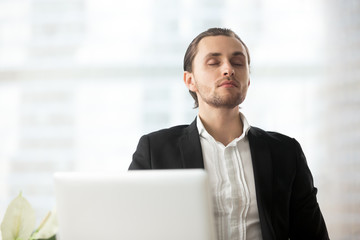 Young calm handsome businessman resting at workplace with eyes closed. Relaxation during workflow, meditation before important meeting, wellness and stress relief at work, maintaining focus concept.