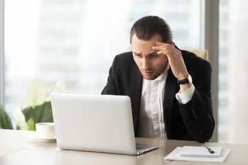 Upset confused businessman looking at laptop screen at workplace in modern office. Stressed young CEO at work desk looking at computer in disbelief about failing report statistics, bad mistake made.