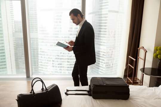 Serious businessman holds guide brochure and calls taxi or room service from hotel apartment. Traveler reads information, looks up address, orders dinner delivery. Trip or vacation concept, side view.