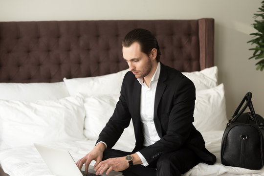 Young businessman sitting on bed beside luggage bag, working on laptop in hotel. Entrepreneur remotely works from hotel room while traveling. Business traveler finalizing work project, browsing web.