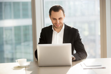Young cheerful business man sitting in modern office, looking at laptop with happy smile, pleased with successful deal, financial report, project results and stock investment profit. Success concept.