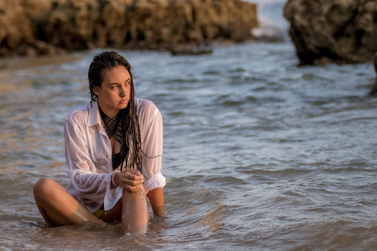Chica joven con camisa blanca sentada en la playa moj&aacute;ndose con las olas del mar al atardecer.