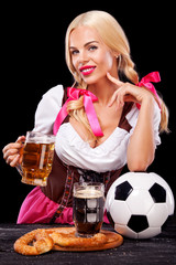 Young sexy Oktoberfest girl - waitress, wearing a traditional Bavarian dress, serving big beer mugs and taking soccer ball on black background.