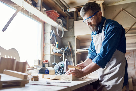 Bearded middle-aged craftsman wearing safety goggles and apron hammering nails into board, interior of messy workshop on background