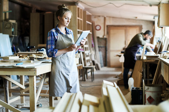 Full length portrait of concentrated young craftswoman in apron standing at spacious workshop and using digital tablet