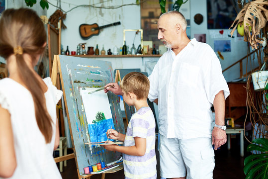 Experienced bearded teacher helping little boy to draw on canvas with watercolors, unrecognizable female pupil looking at them with interest, interior of art studio on background