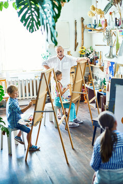 Group of gifted children sitting in front of easels and creating still-life paintings, their senior teacher keeping eye on them, interior of spacious art studio on background