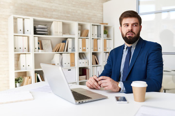 Portrait of successful  bearded businessman looking away thinking while working with laptop  in modern office