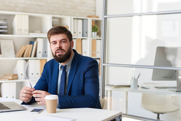 Portrait of successful  bearded businessman looking away thoughtfully while working at desk in modern office