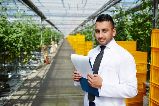 Waist-up portrait of handsome young specialist looking at camera with confident smile while carrying out inspection at spacious modern greenhouse