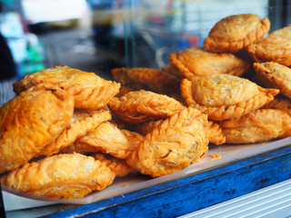 Small handmade folded deep fried crispy golden brown curry puff pie piled up in transparent glass cabinet of Thai local traditional street food stall cart