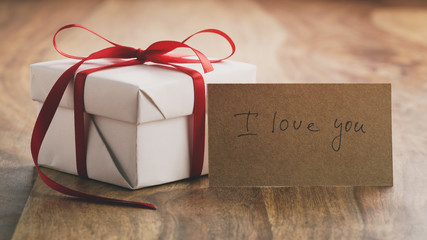 white gift box with thin red ribbon bow on old wood table with i love you paper card