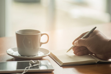 Businessman hand holding a pen writing on paper notebook with coffee cup and smart phone on office table.